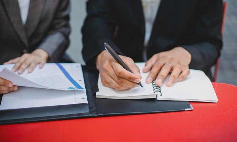 Two professionals in suits writing notes and reviewing documents at an outdoor meeting table.