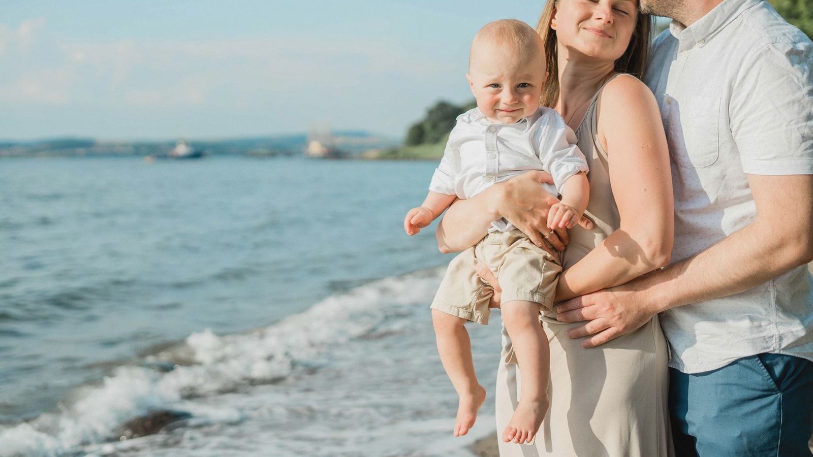 A joyful family moment captured by the sea, with a couple holding their smiling baby on a sunny day.