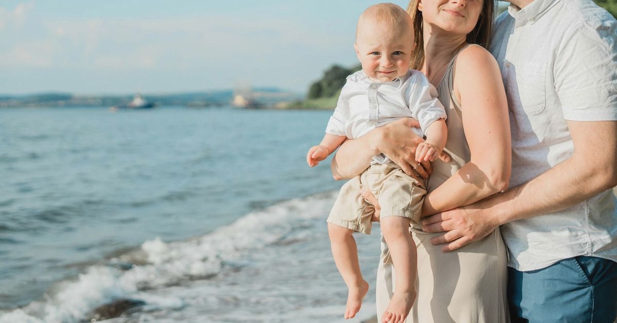 A joyful family moment captured by the sea, with a couple holding their smiling baby on a sunny day.