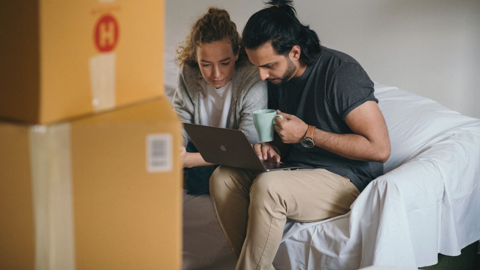 Ethnic man with cup of hot drink typing on netbook while sitting near pensive girlfriend in casual wear on cozy sofa near pile of boxes preparing to relocate
