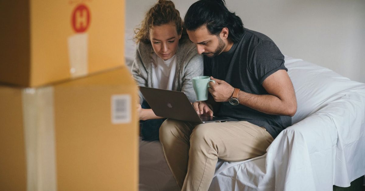 Ethnic man with cup of hot drink typing on netbook while sitting near pensive girlfriend in casual wear on cozy sofa near pile of boxes preparing to relocate