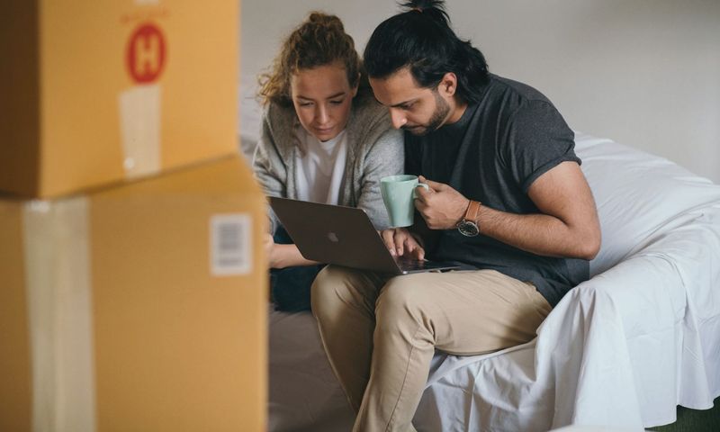 Ethnic man with cup of hot drink typing on netbook while sitting near pensive girlfriend in casual wear on cozy sofa near pile of boxes preparing to relocate