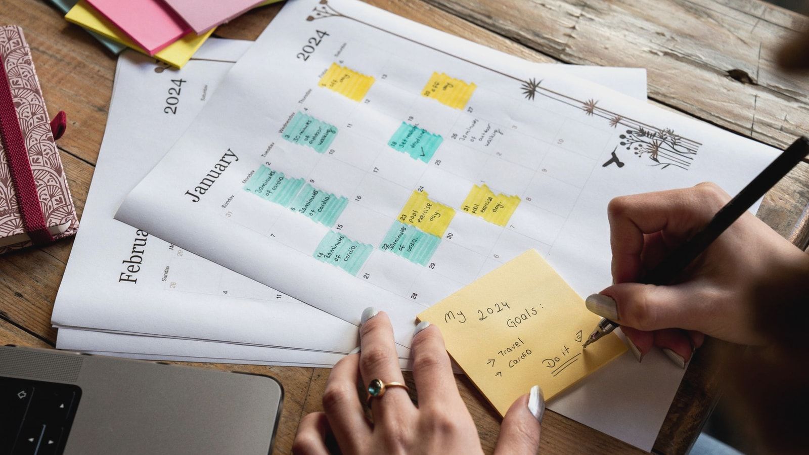 Close-up of a person writing new year's goals on a sticky note with a 2024 calendar in the background.