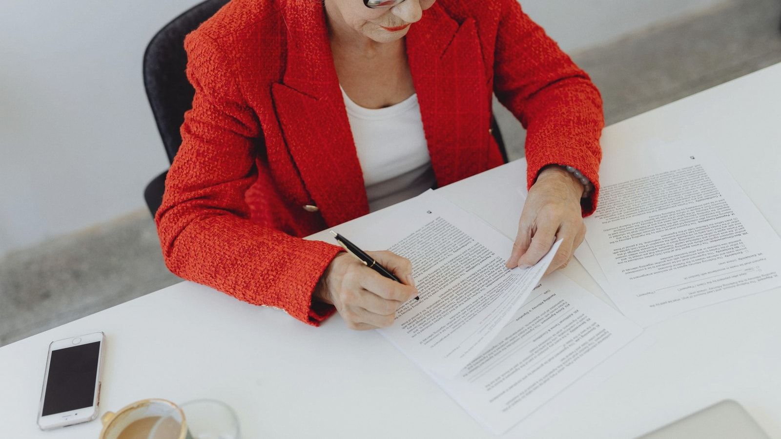 Elderly businesswoman in red jacket signing papers at office desk, showcasing leadership.