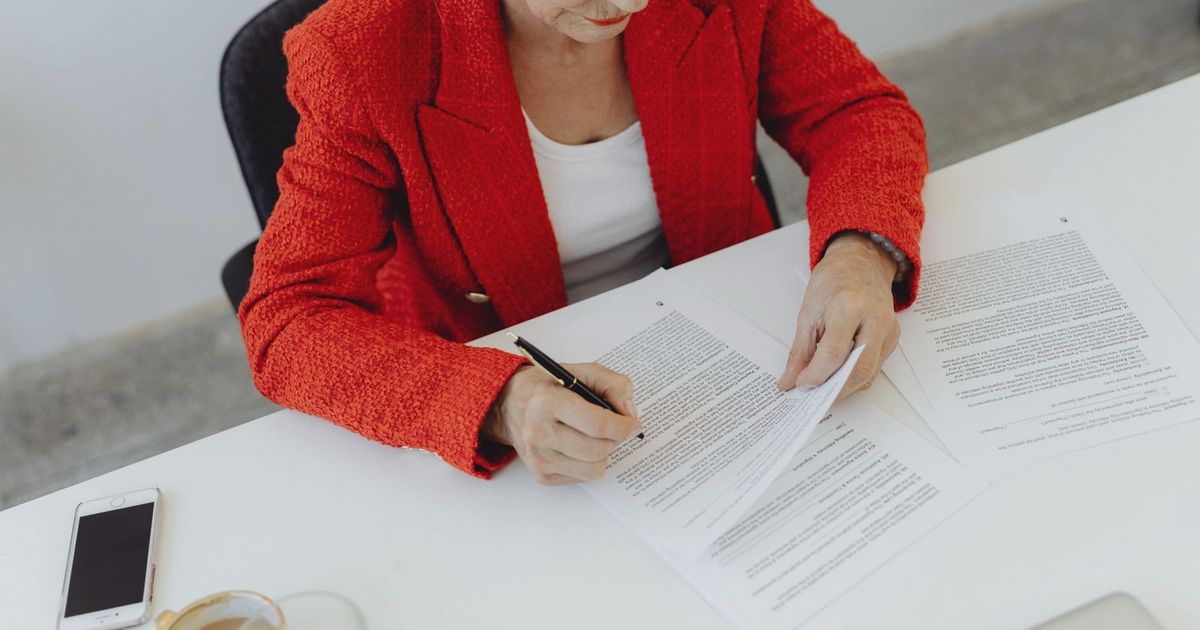 Elderly businesswoman in red jacket signing papers at office desk, showcasing leadership.