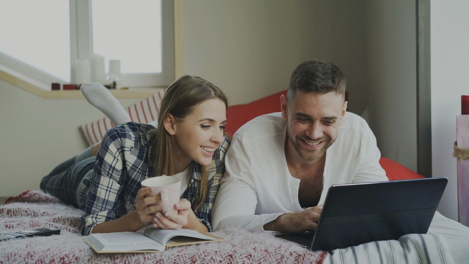Smiling man and woman talking and lying in bed reading book and using laptop computer in the morning
