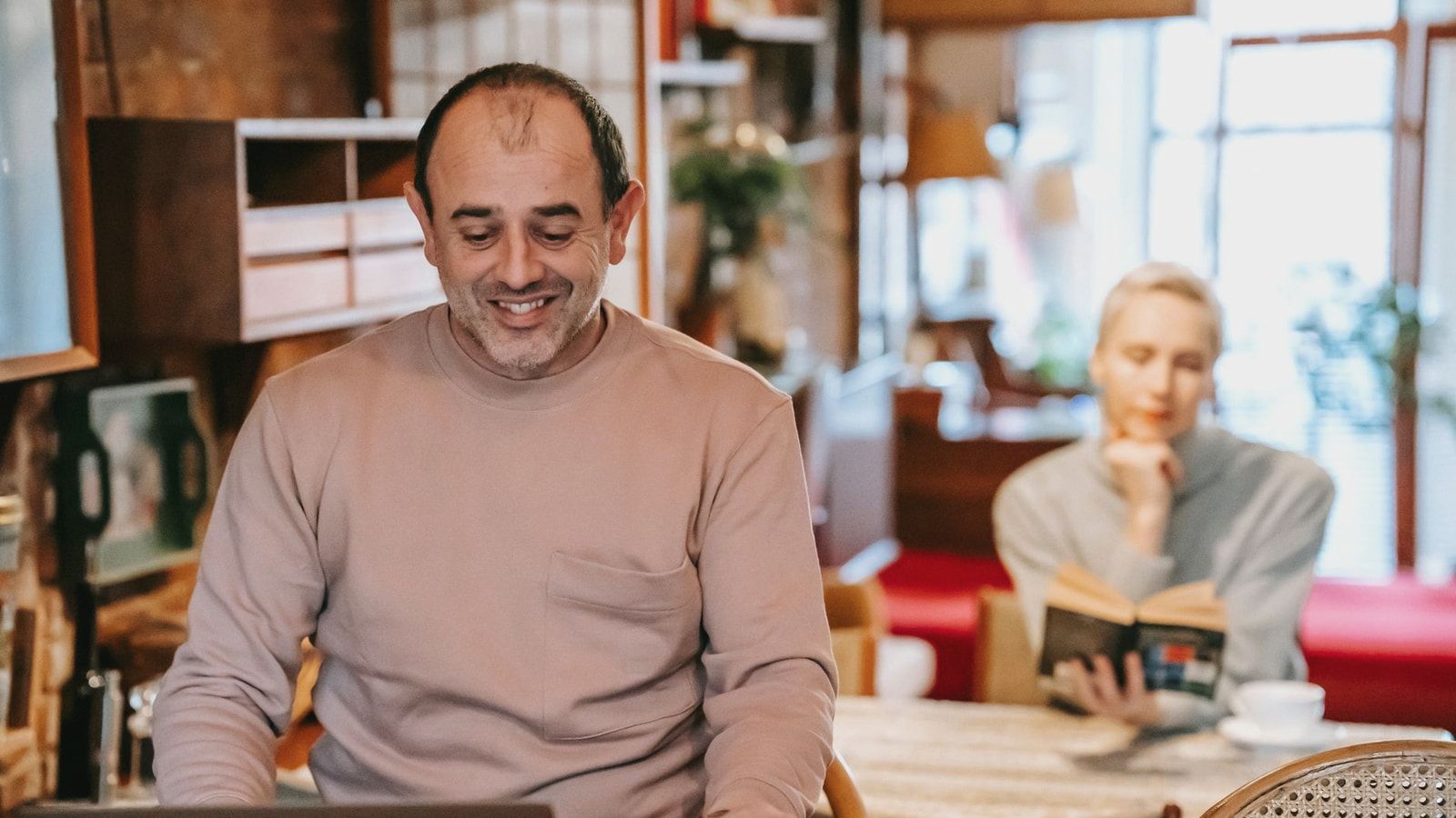 Positive middle aged ethnic male freelancer drinking coffee and working remotely in laptop near focused wife reading book at home