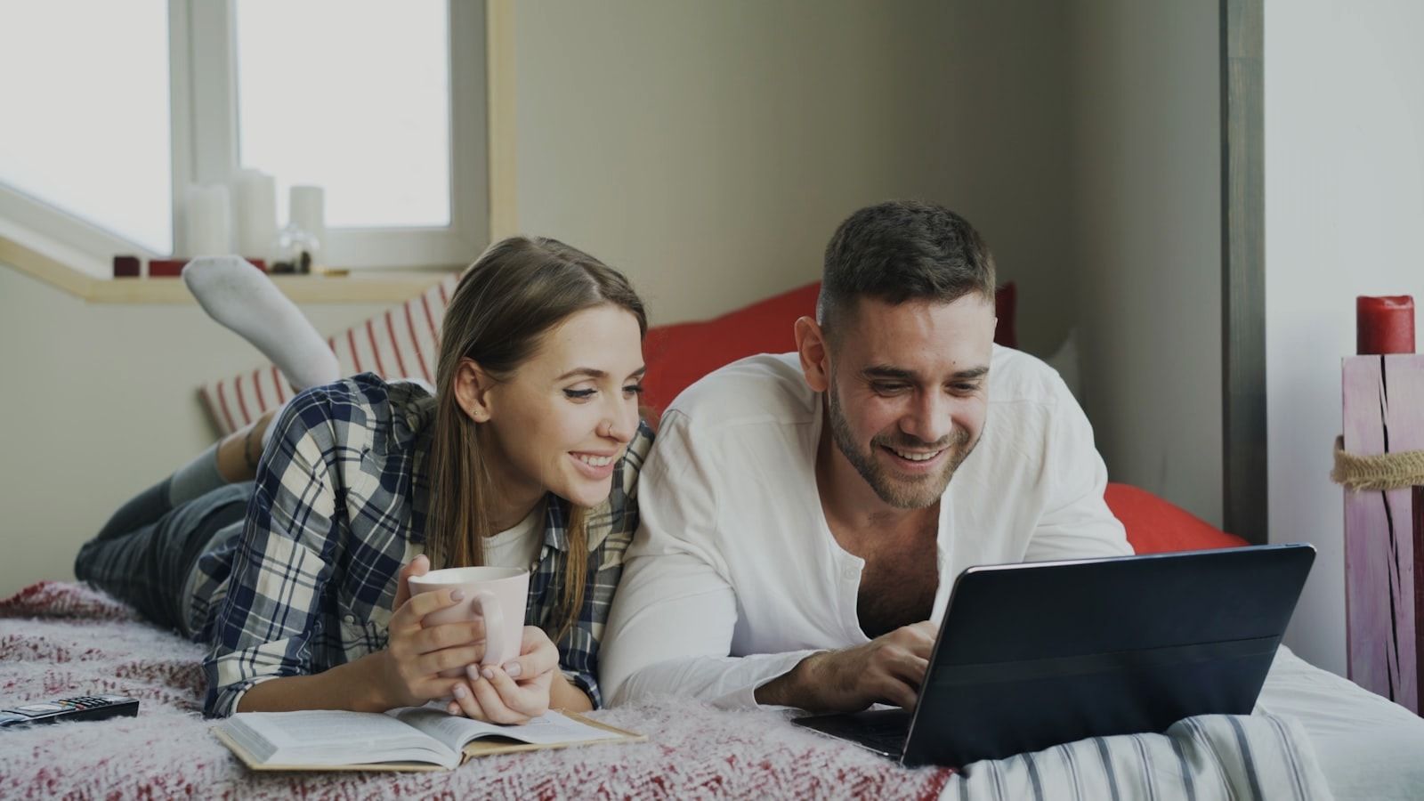 Smiling man and woman talking and lying in bed reading book and using laptop computer in the morning