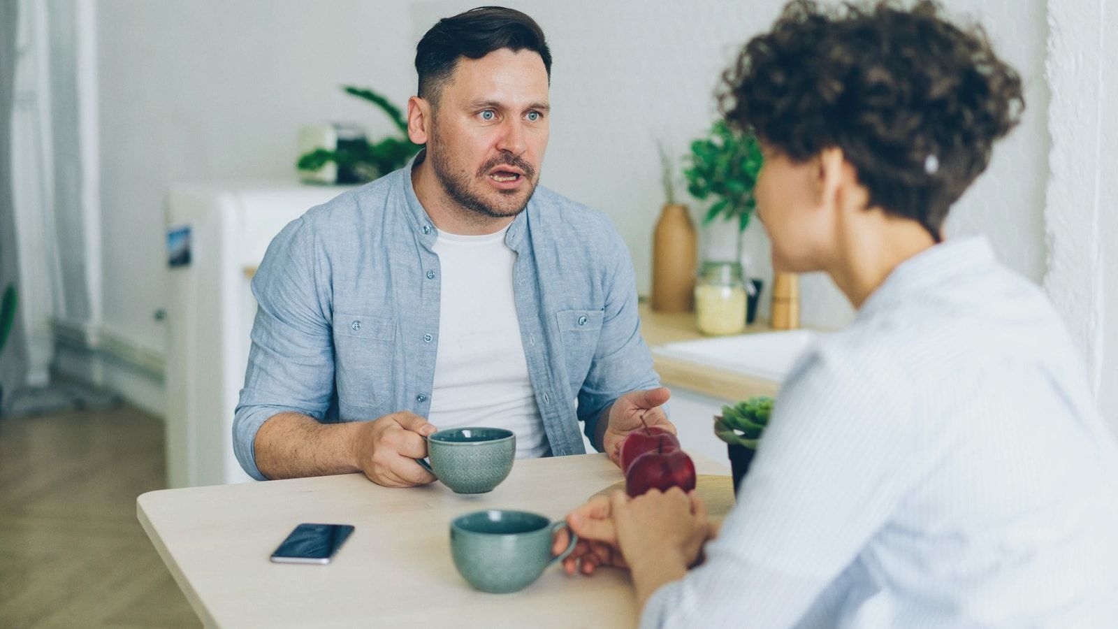 Couple having a serious conversation at the kitchen table with coffee cups.
