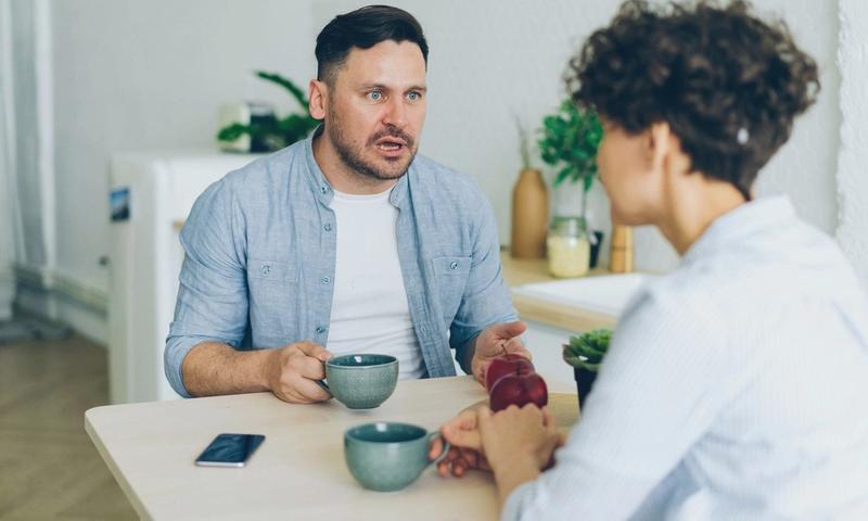 Couple having a serious conversation at the kitchen table with coffee cups.