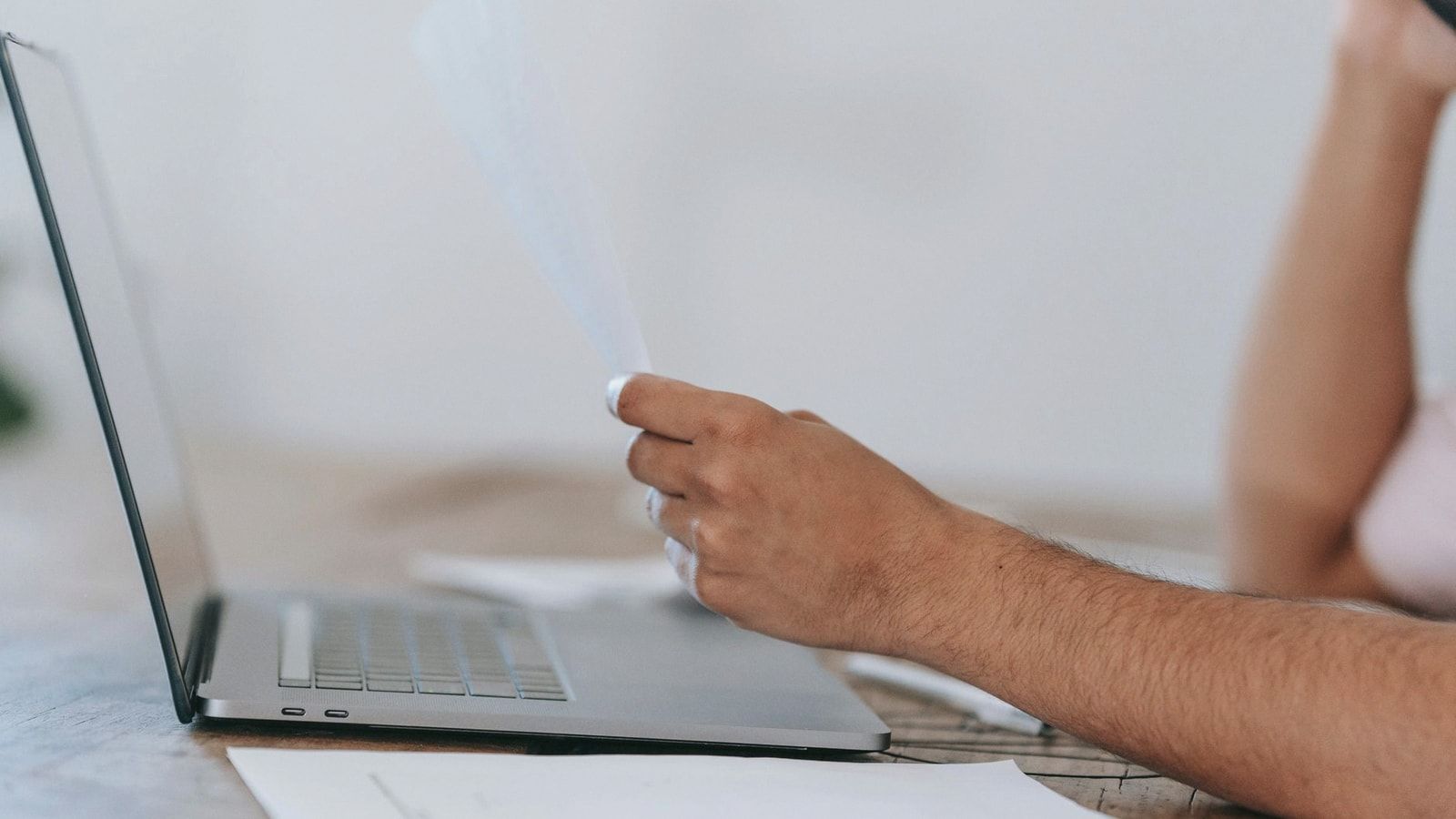Crop anonymous male reading document and talking on mobile phone while sitting at desk with laptop