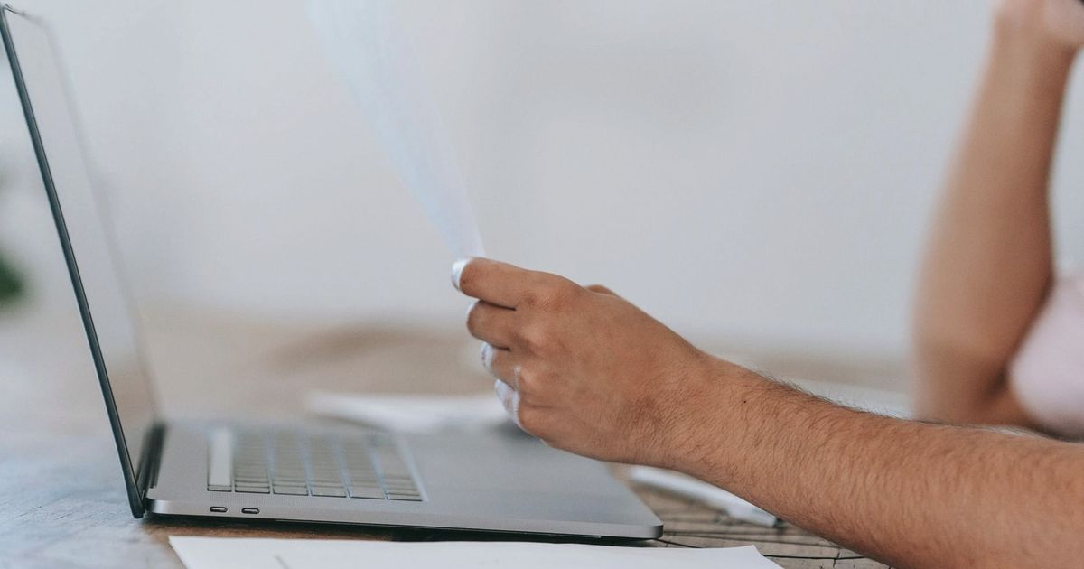 Crop anonymous male reading document and talking on mobile phone while sitting at desk with laptop
