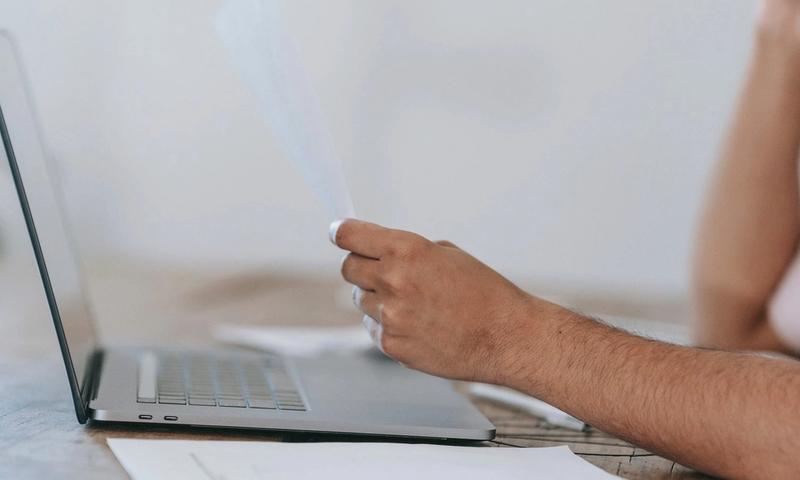 Crop anonymous male reading document and talking on mobile phone while sitting at desk with laptop