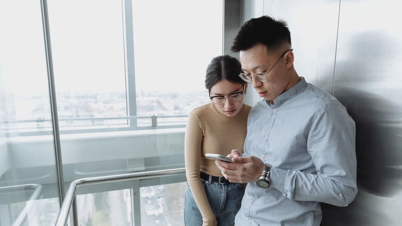 Asian man and woman looking at smartphone in modern glass elevator with city view.