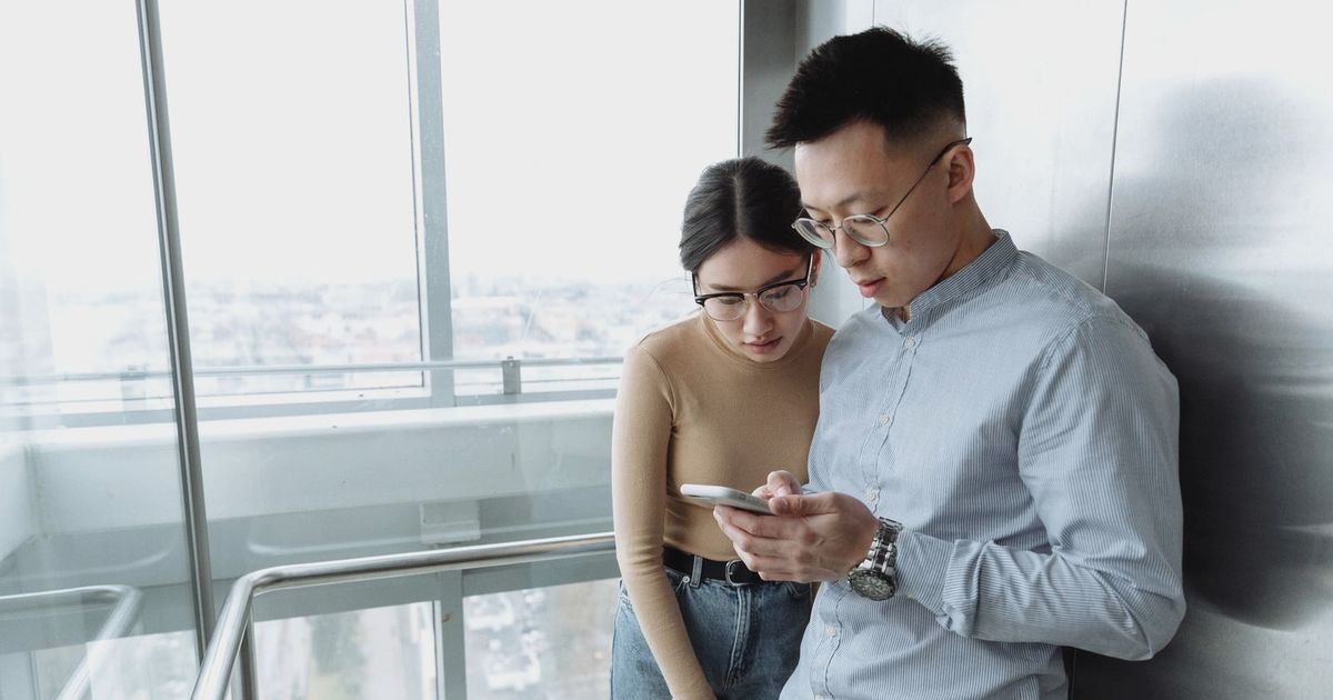 Asian man and woman looking at smartphone in modern glass elevator with city view.