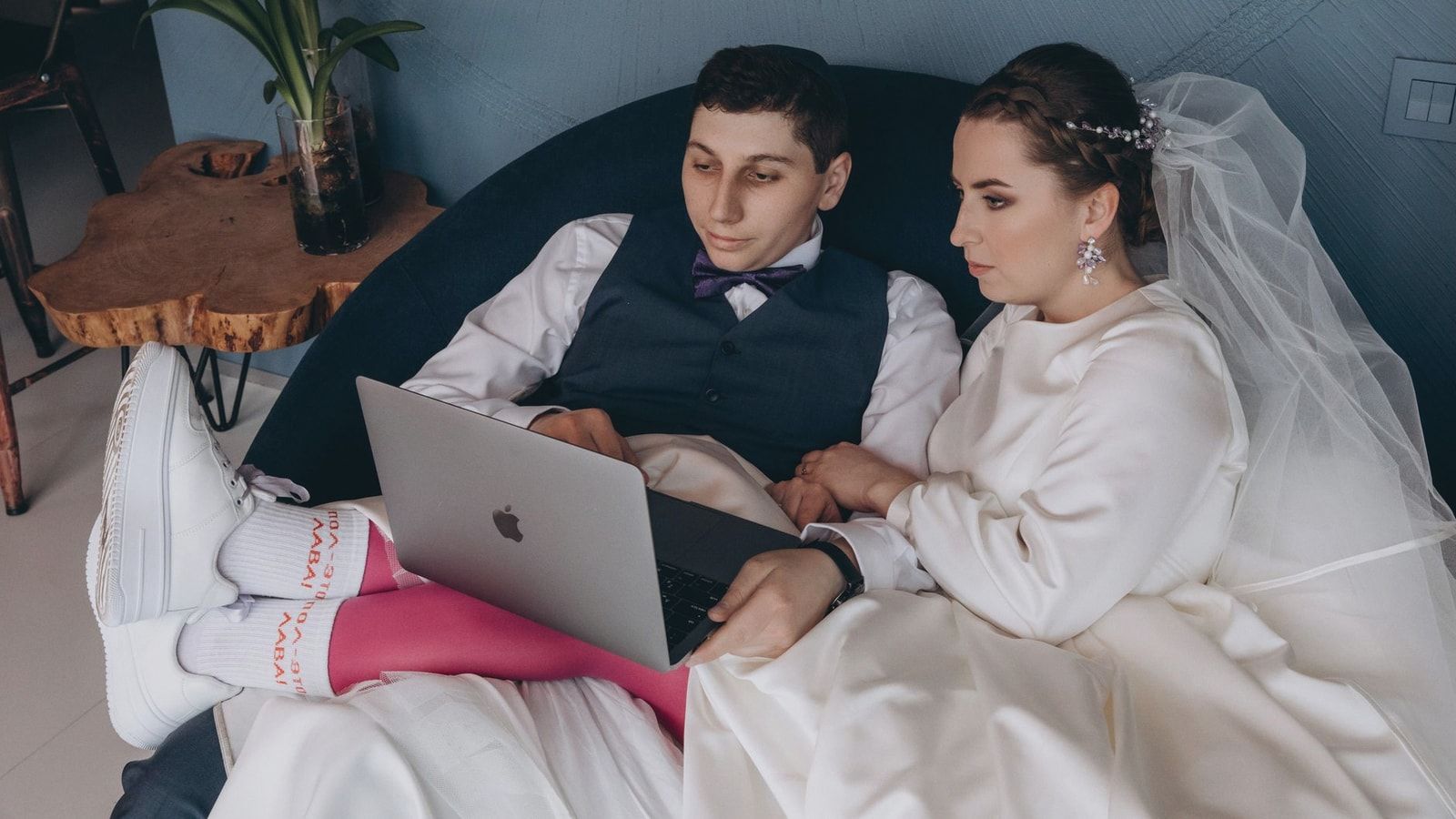 A bride and groom sitting indoors, browsing a laptop together in wedding attire.