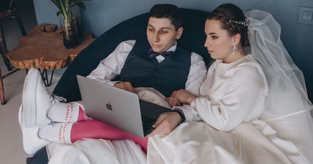A bride and groom sitting indoors, browsing a laptop together in wedding attire.