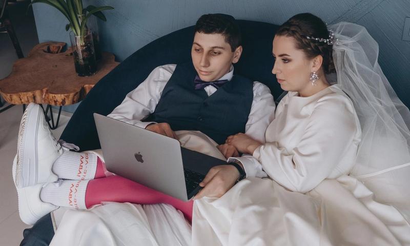 A bride and groom sitting indoors, browsing a laptop together in wedding attire.