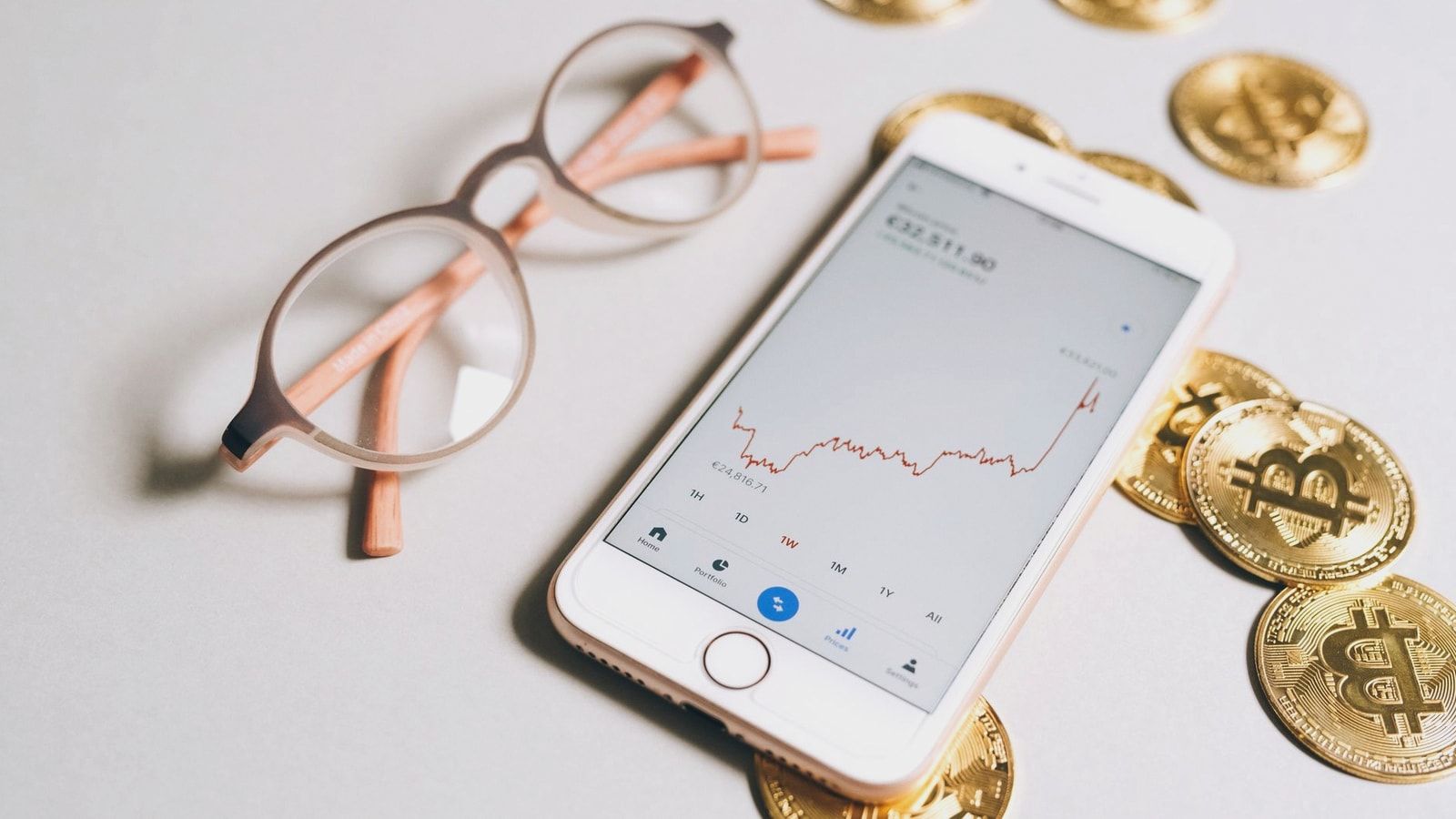 Smartphone displaying cryptocurrency graph alongside bitcoin coins and eyeglasses.