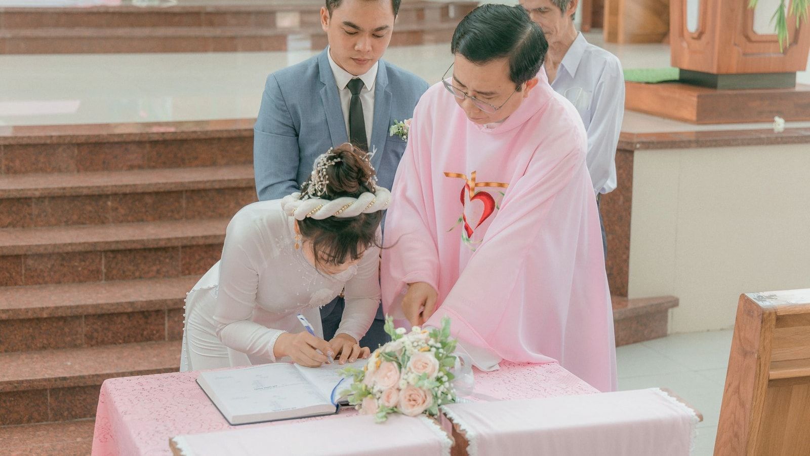 Bride and groom signing marriage documents with priest during church ceremony.
