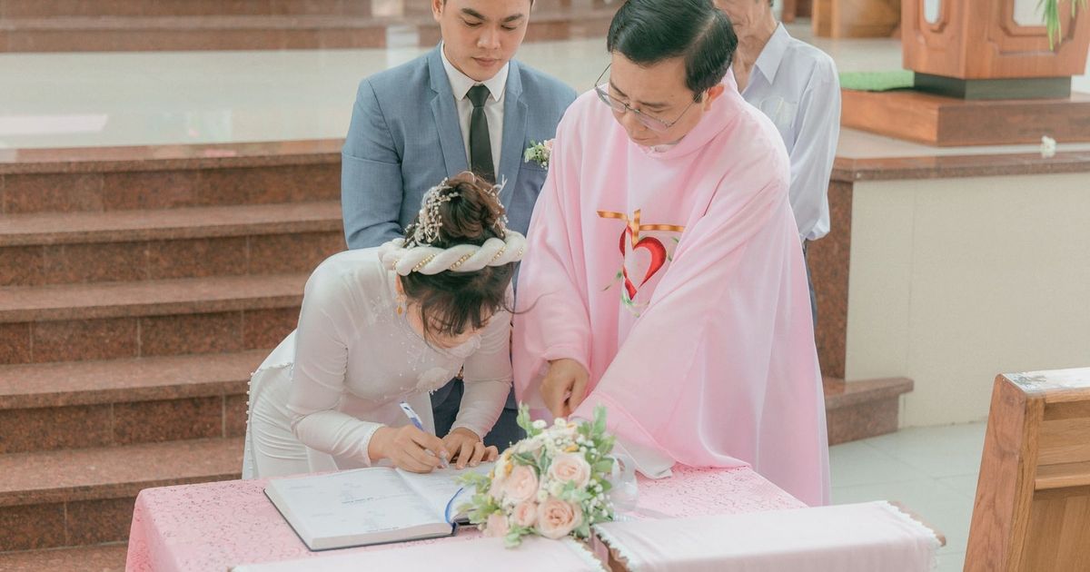 Bride and groom signing marriage documents with priest during church ceremony.
