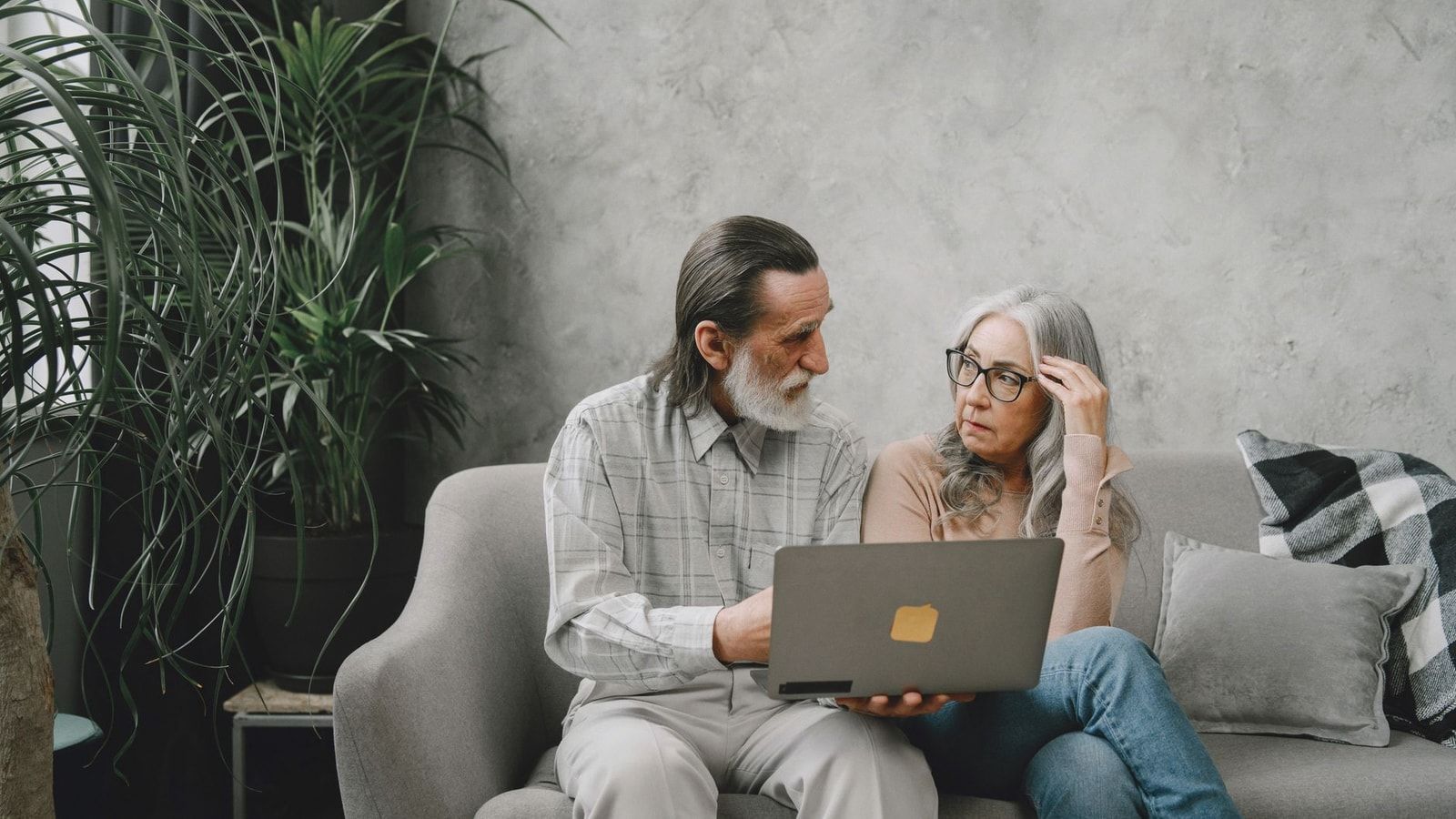 Elderly couple discussing while using laptop at home on sofa, surrounded by plants.
