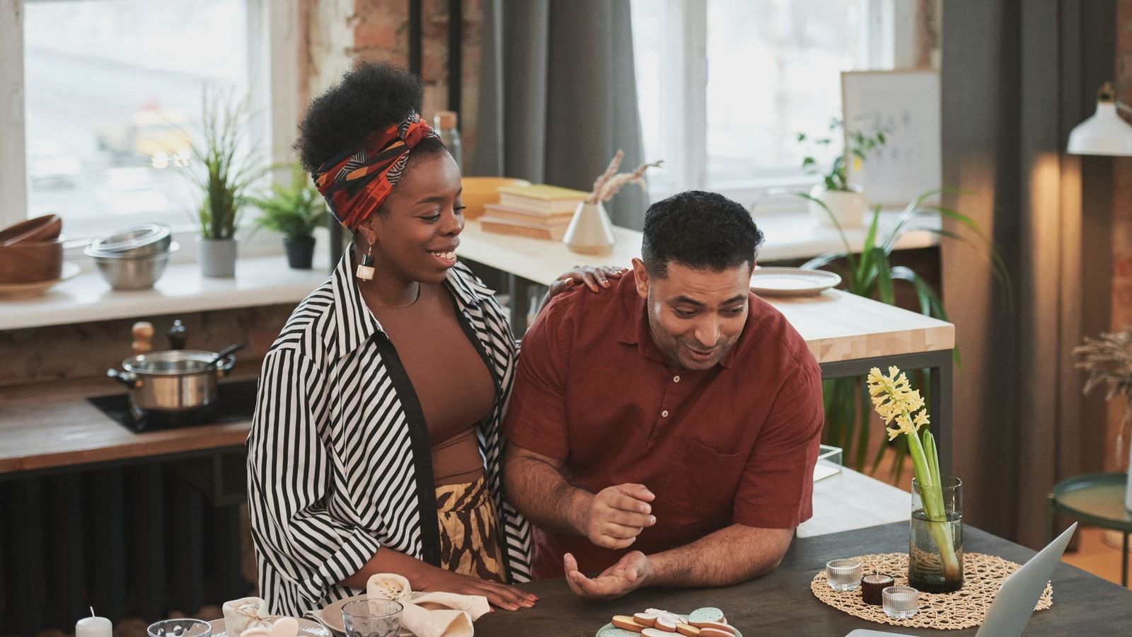 Smiling couple baking cookies together in a cozy kitchen using a laptop for recipe checking.