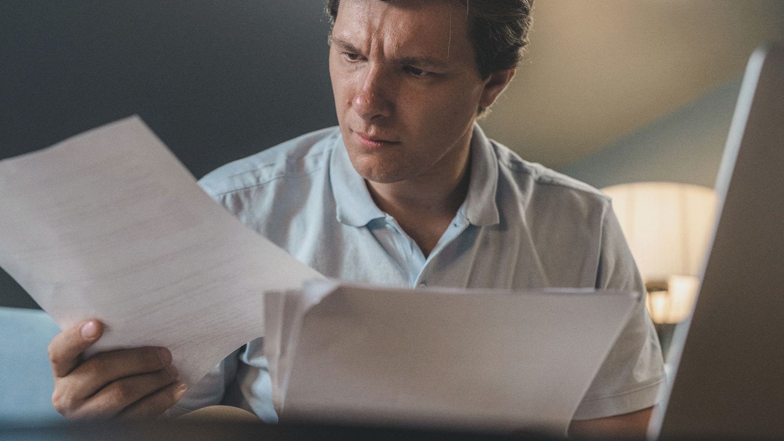 A man intently examines papers, seated indoors under warm lighting, focusing on his work.