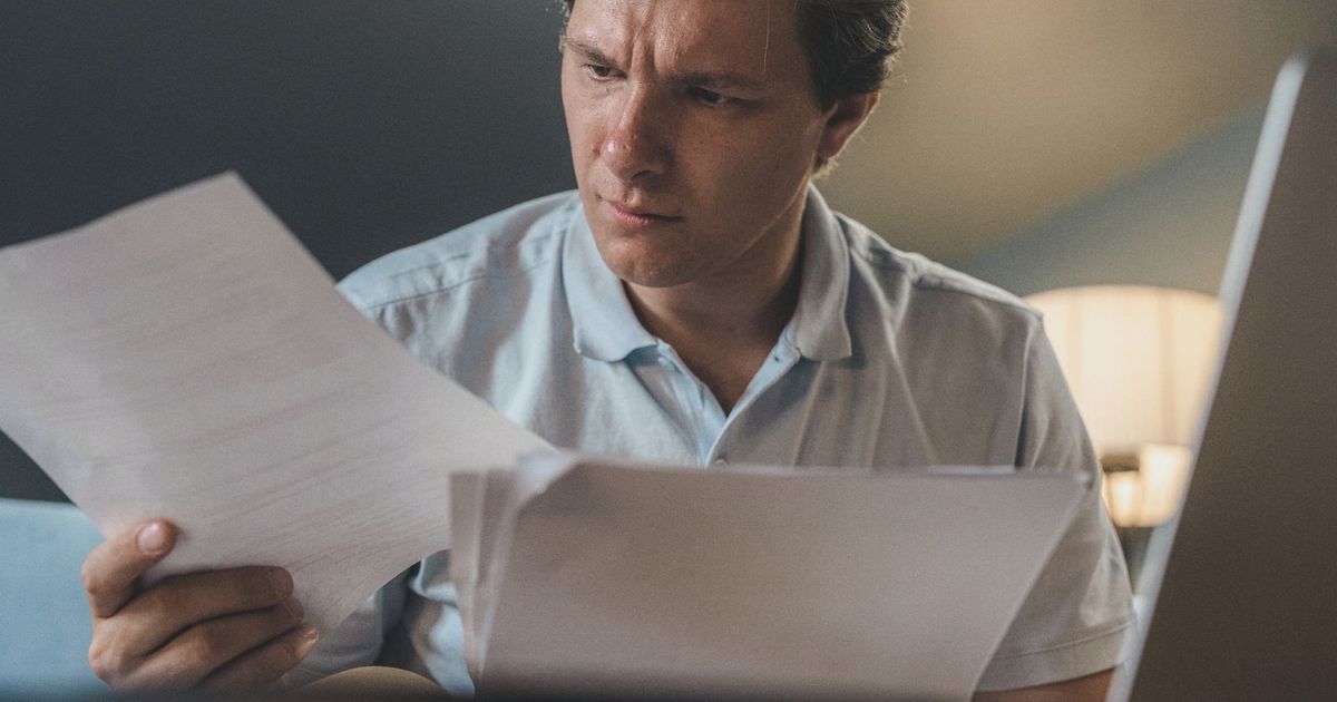 A man intently examines papers, seated indoors under warm lighting, focusing on his work.