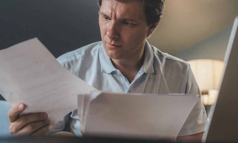 A man intently examines papers, seated indoors under warm lighting, focusing on his work.