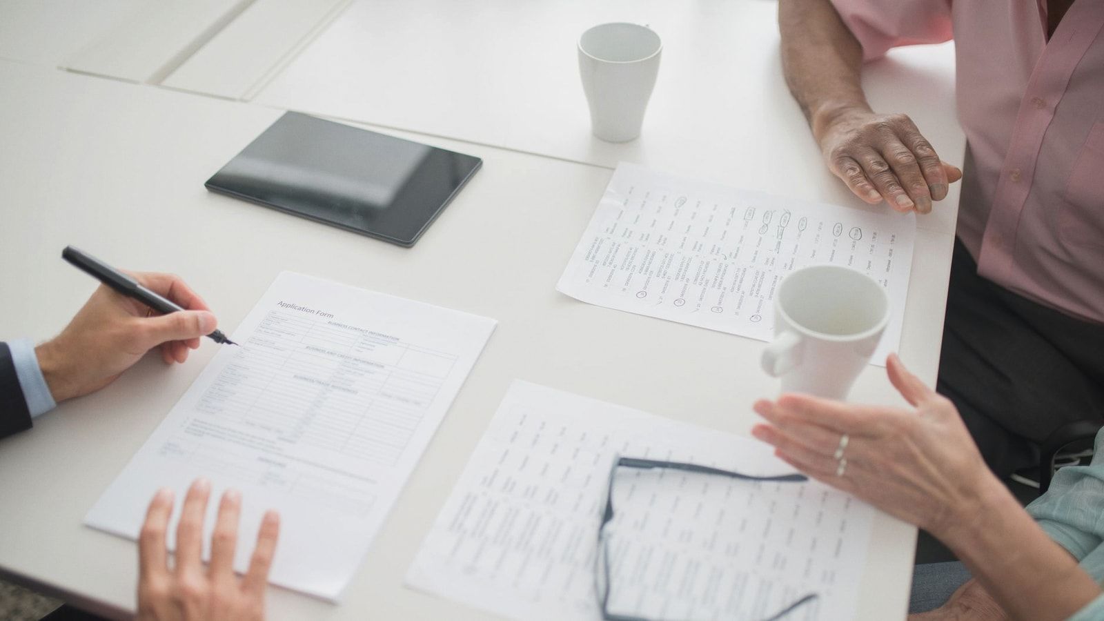 Hands writing on documents in a business meeting setup with coffee cups and tablet.