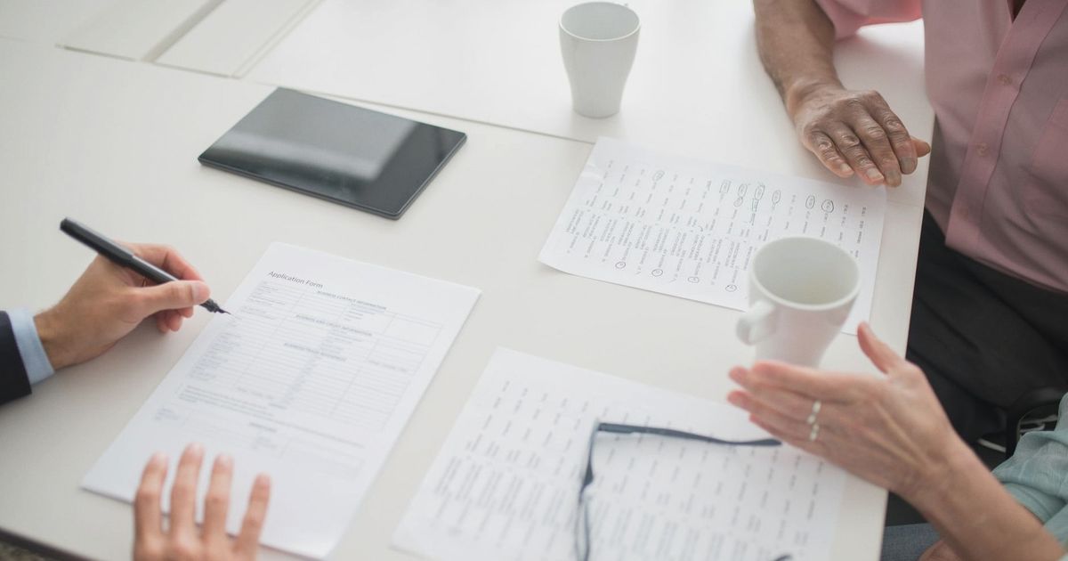 Hands writing on documents in a business meeting setup with coffee cups and tablet.