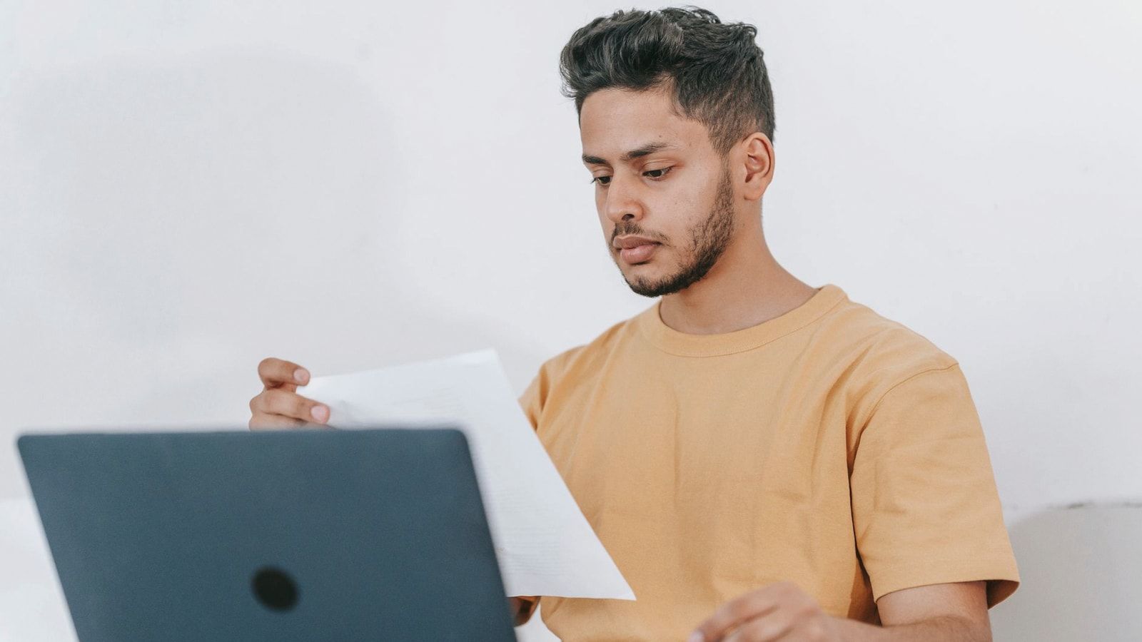 Young bearded Hispanic male university student reading information on paper while studying online via laptop