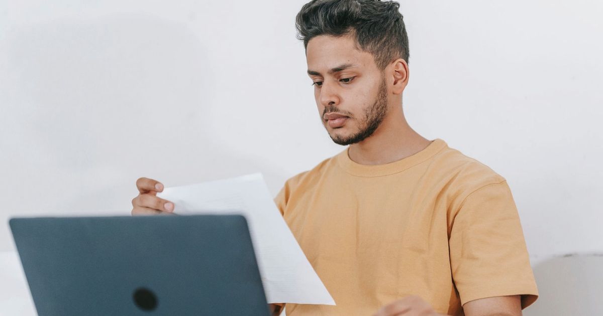 Young bearded Hispanic male university student reading information on paper while studying online via laptop
