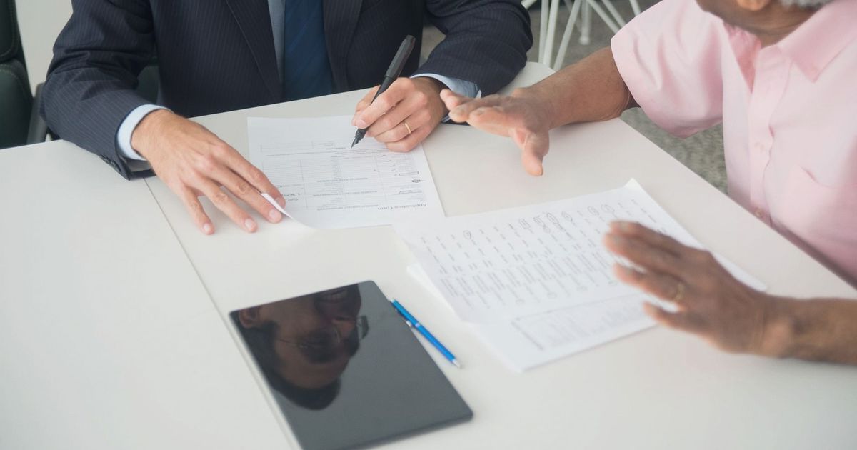 A business meeting between an elderly client and a consultant discussing documents at an office table.