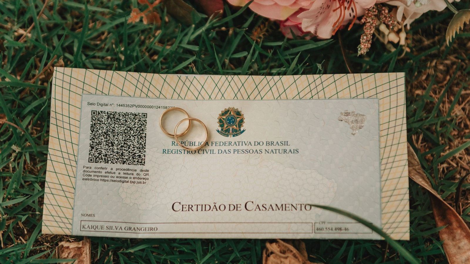 Close-up of a Brazilian marriage certificate with wedding rings and flowers on grass.
