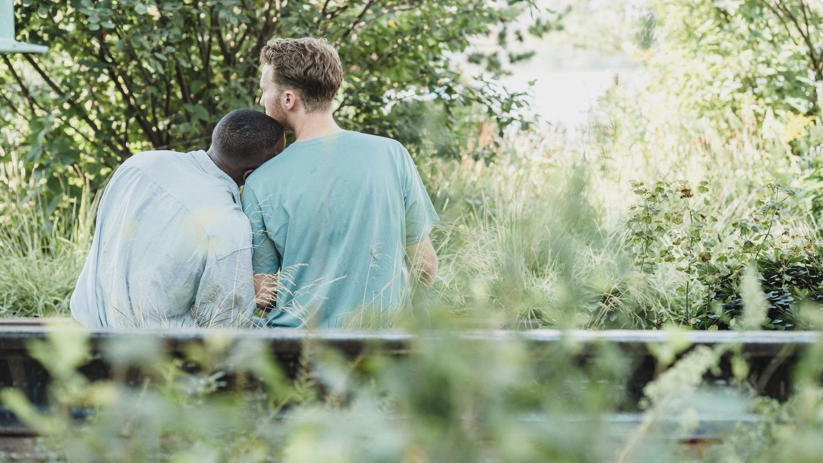 Two men sitting together on a bench in a lush park, showcasing affection and companionship.