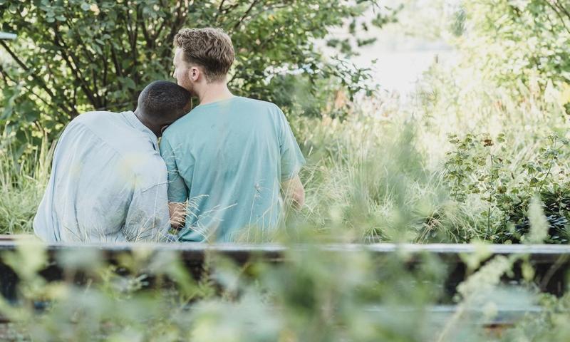 Two men sitting together on a bench in a lush park, showcasing affection and companionship.