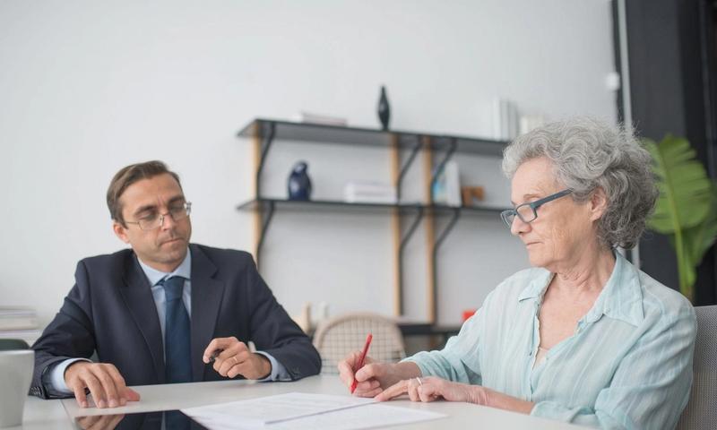Elderly woman signing paperwork in modern office with consultant.