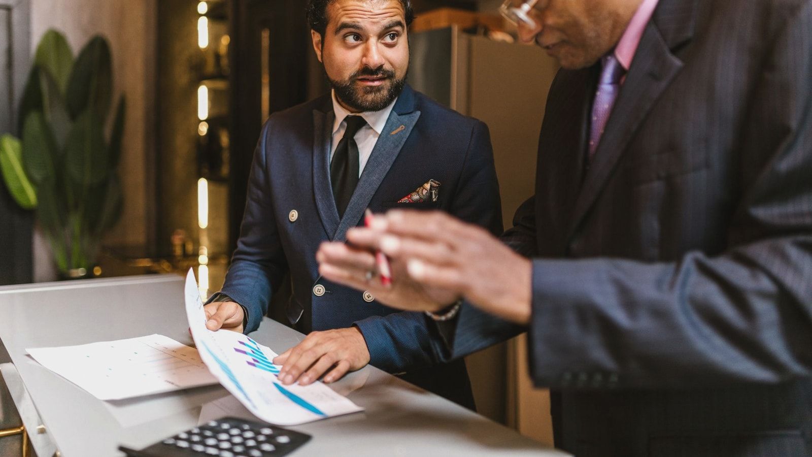 Two men in suits discuss financial documents with graphs indoors.