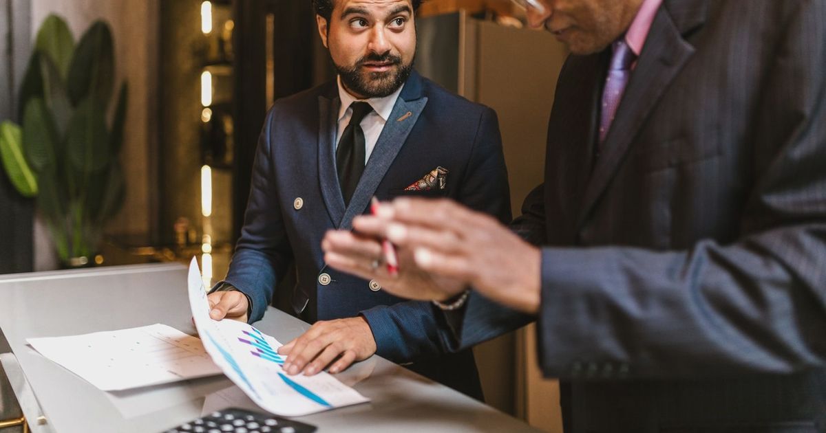 Two men in suits discuss financial documents with graphs indoors.