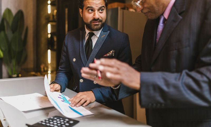 Two men in suits discuss financial documents with graphs indoors.