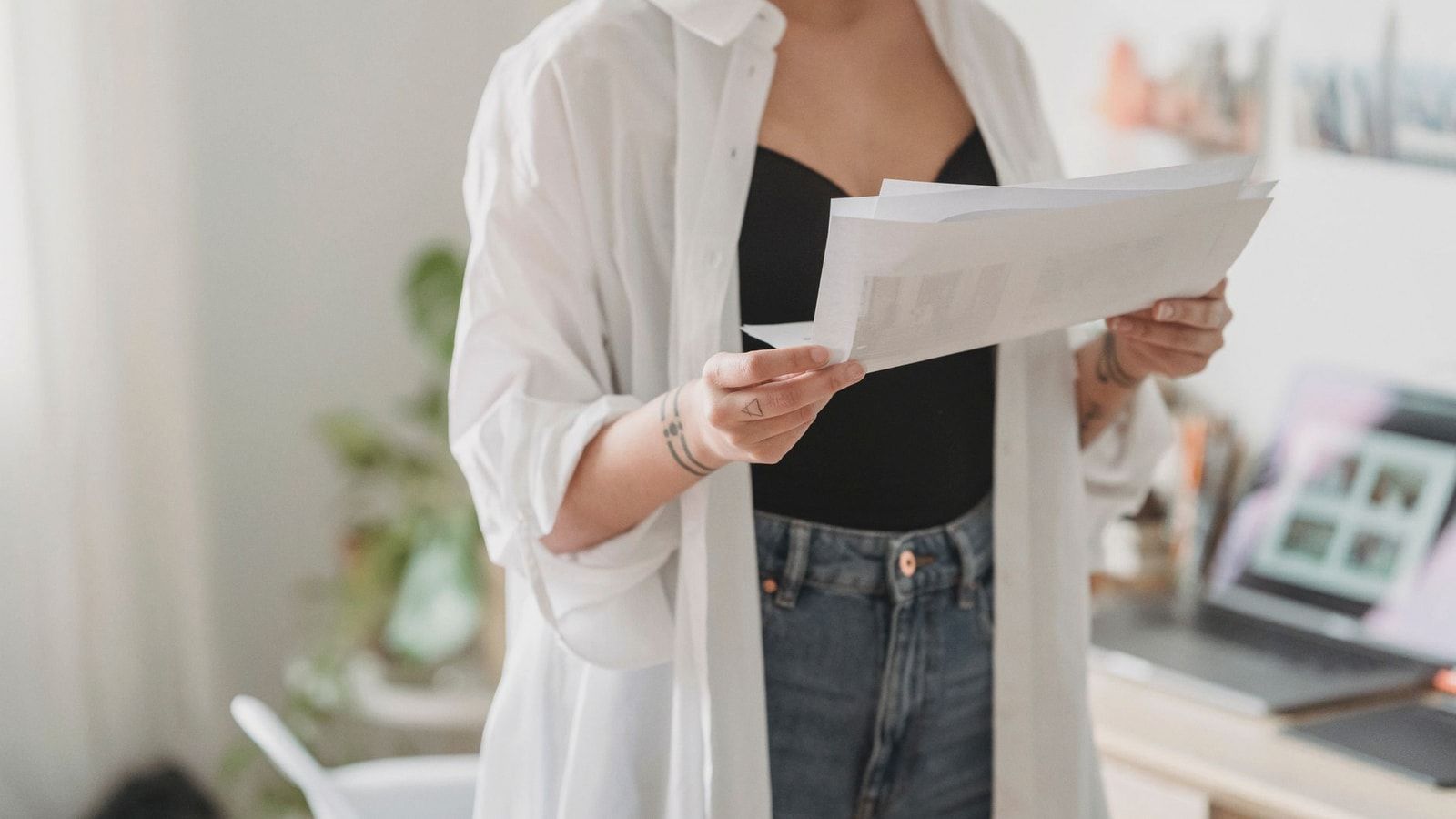 Crop young thoughtful woman with tattoos and paper sheets working against laptop at home in daytime