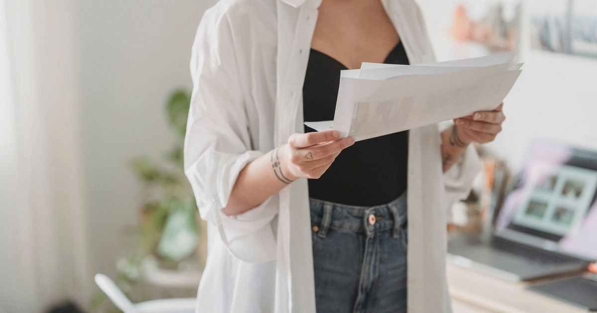Crop young thoughtful woman with tattoos and paper sheets working against laptop at home in daytime