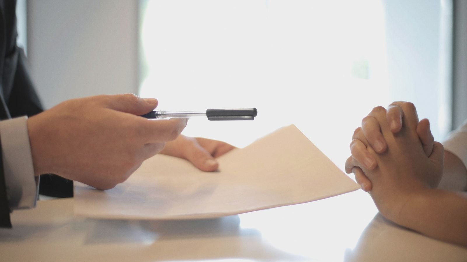 Side view of faceless formal man giving pen and paper to focused female with clenched hands at table on meeting