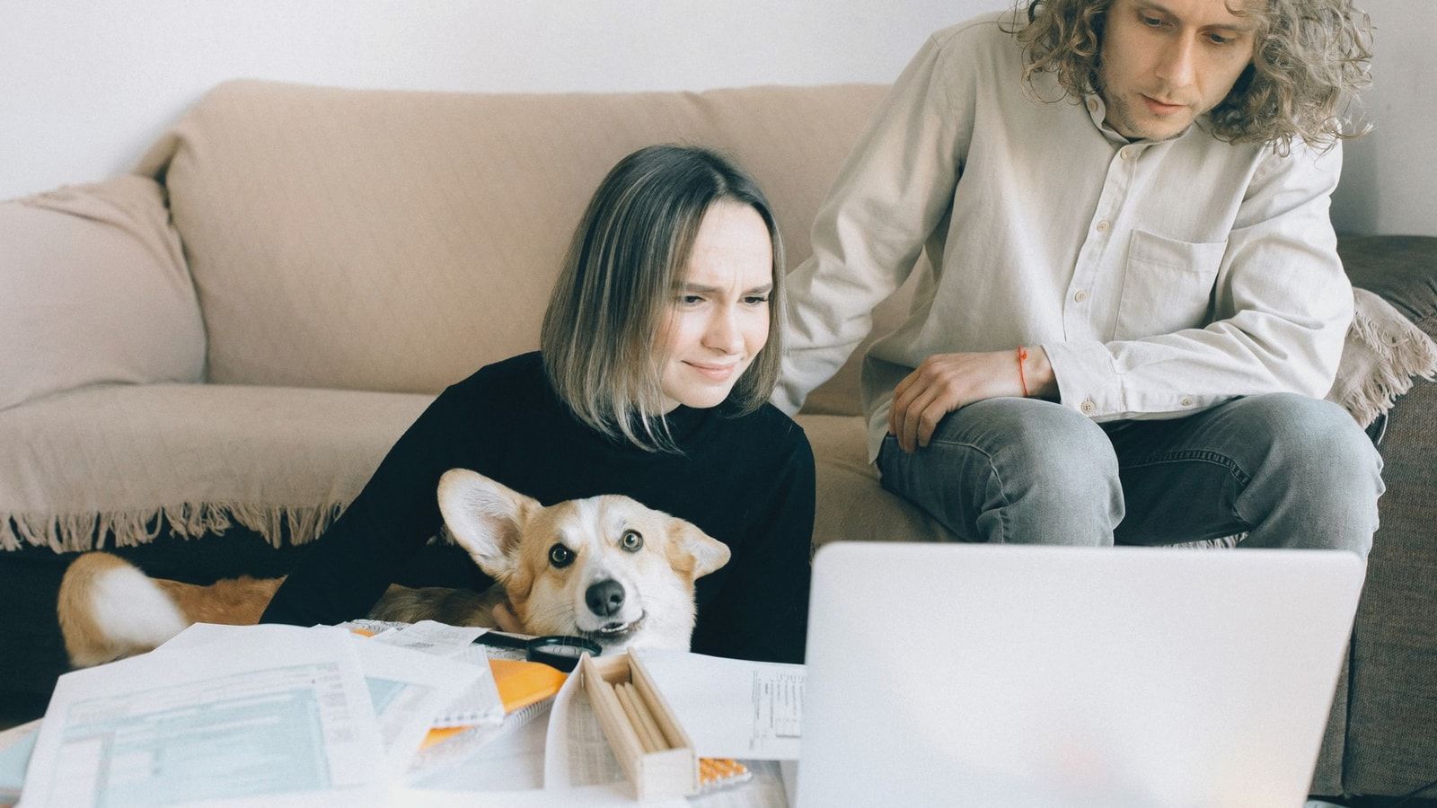 A couple looking at a laptop while sitting on a couch with their pet Corgi amidst paperwork indoors.