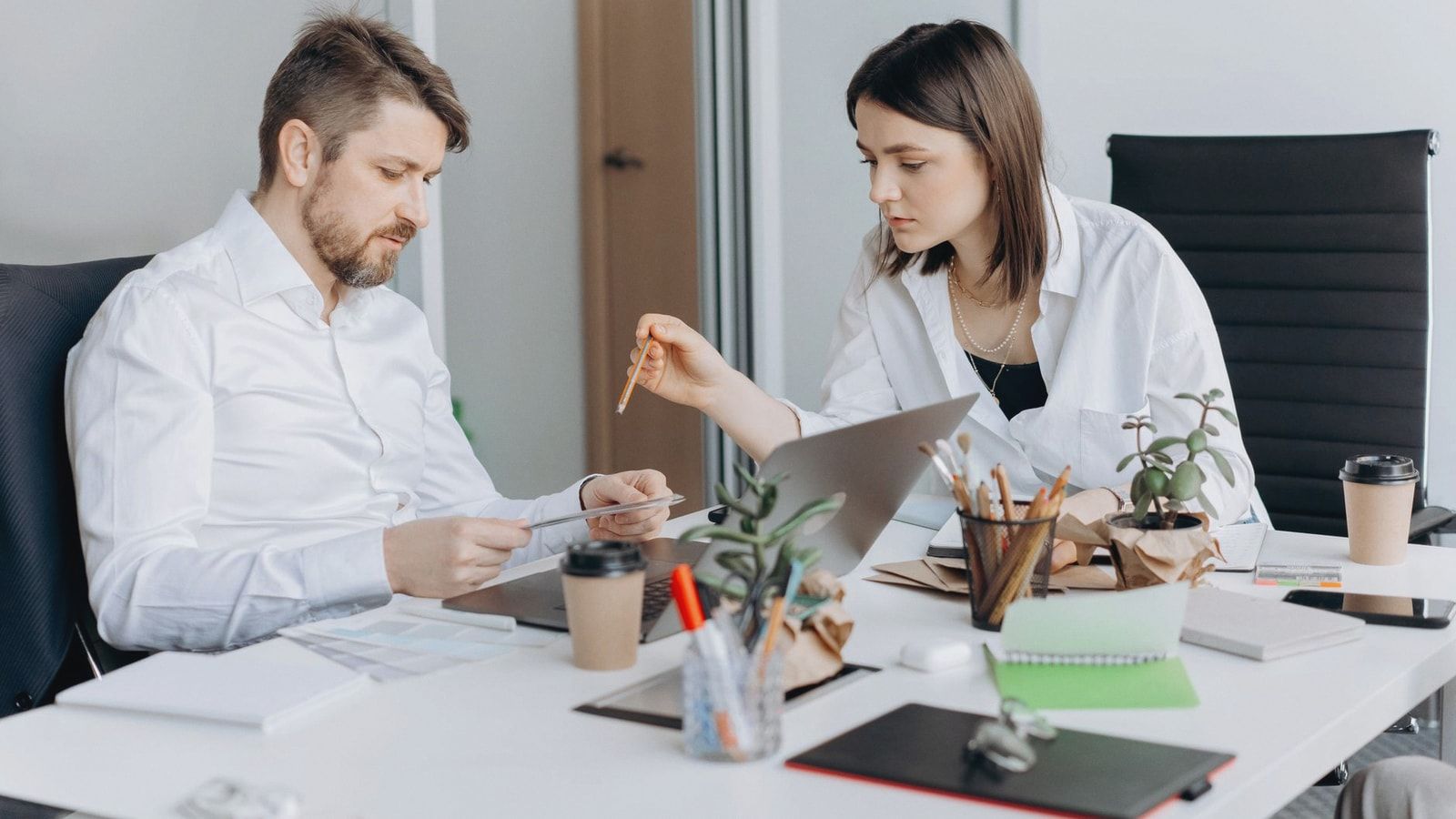 Two colleagues discussing work at a desk with laptops and coffee in a modern office setting.