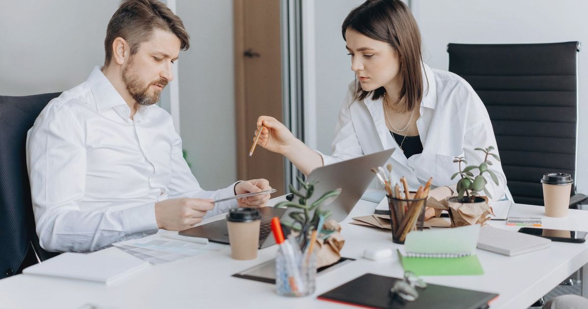 Two colleagues discussing work at a desk with laptops and coffee in a modern office setting.