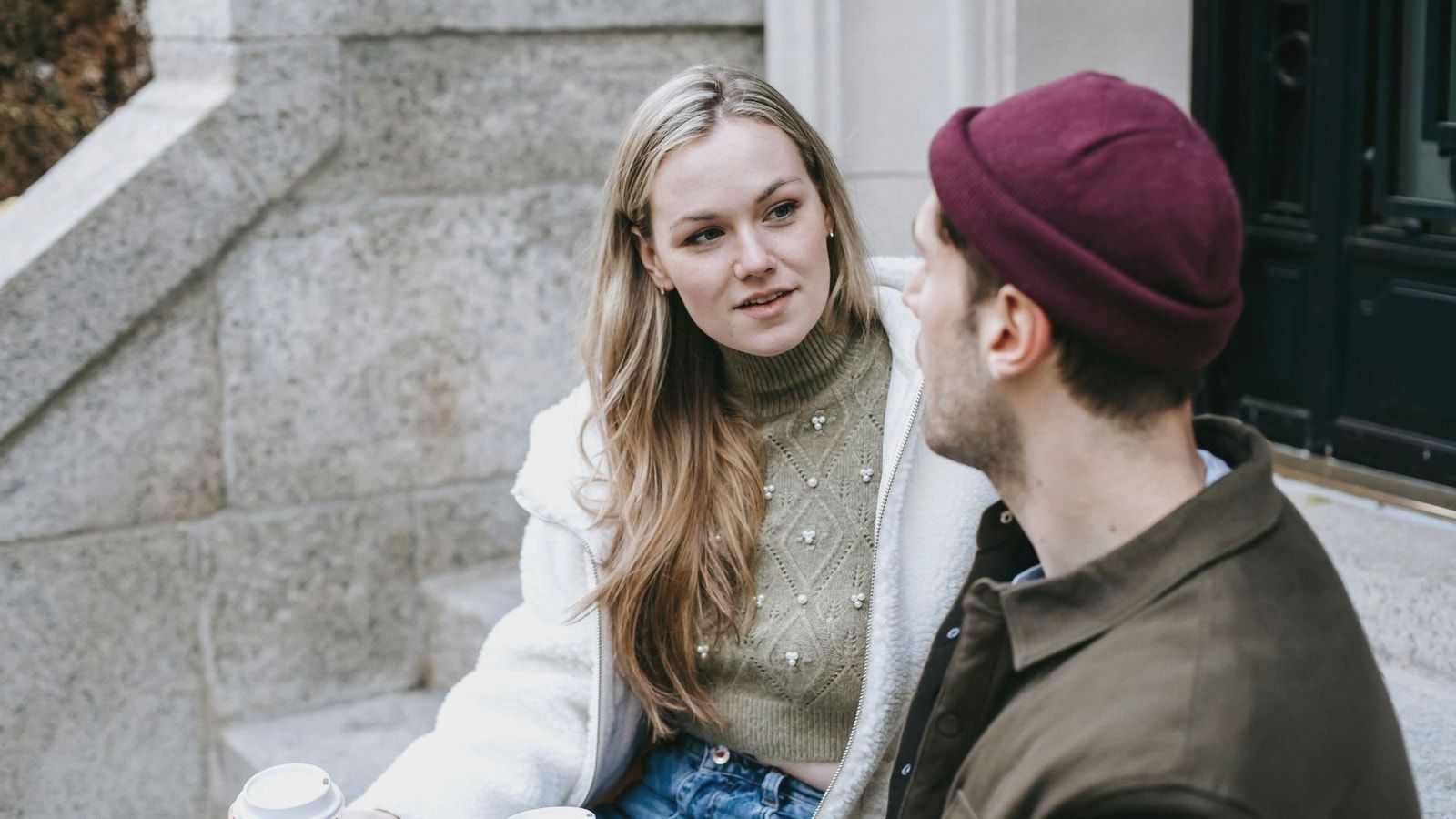 Man and woman talking sitting on steps outside house and having hot beverage in paper cups