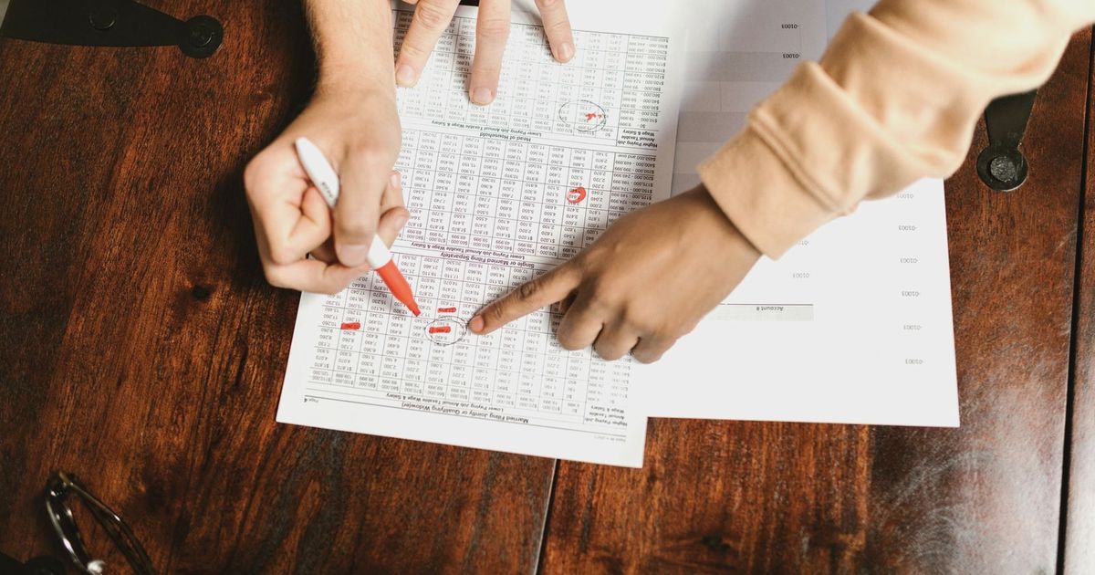 Two hands reviewing and marking financial documents with a red pen on a wooden table, top view.
